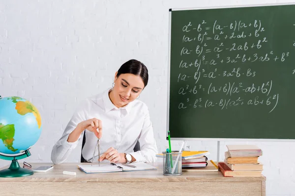 Smiling teacher holding drawing compass near books and notebooks on table — Stock Photo