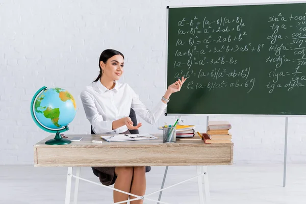 Smiling teacher pointing at chalkboard near globe and books in classroom — Stock Photo