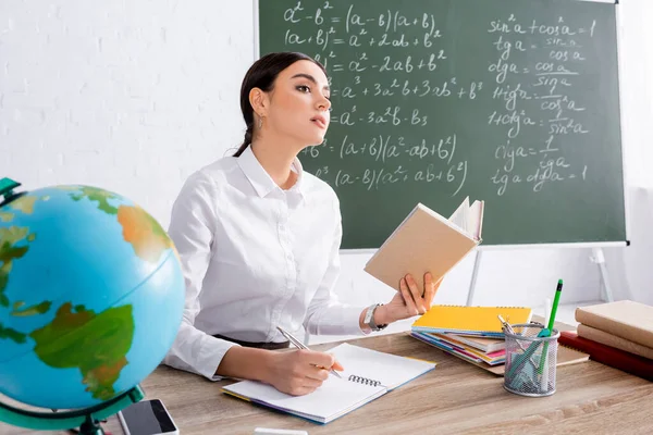 Teacher holding book and writing on notebook near globe in classroom — Stock Photo