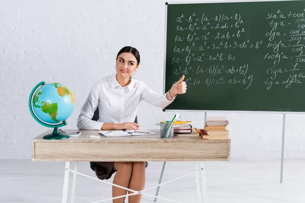 Teacher showing thumb up near smartphone, globe and books in classroom — Stock Photo