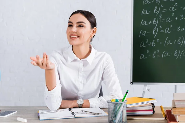 Smiling teacher sitting near books, smartphone and chalk in classroom — Stock Photo