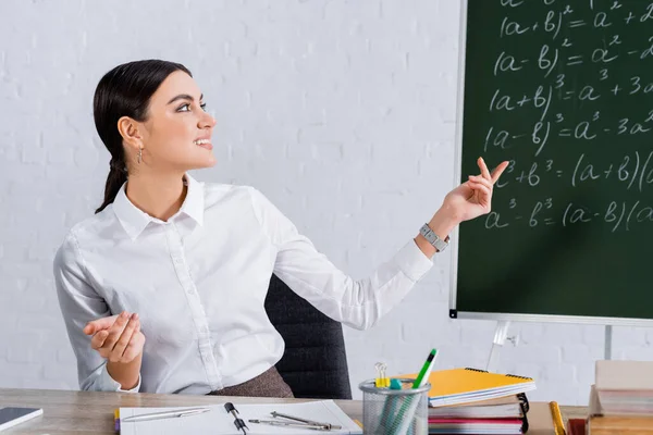 Teacher pointing at chalkboard near notebooks and smartphone on table — Stock Photo