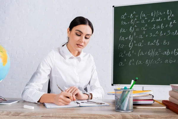 Young teacher writing on notebook near drawing compass and books in classroom — Stock Photo