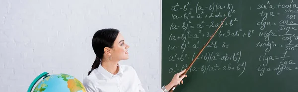 Улыбающийся учитель, указывающий на математические уравнения на доске возле земного шара, баннер — стоковое фото