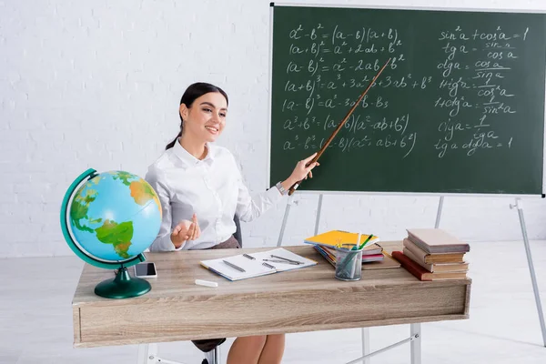 Cheerful teacher with pointer sitting near chalkboard and globe during lesson — Stock Photo