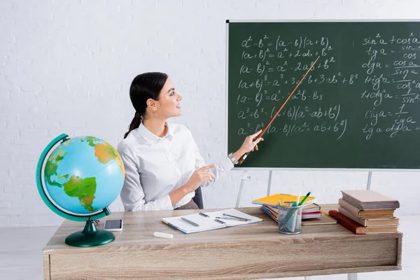 Smiling teacher pointing at chalkboard with equations near books and globe in school — Stock Photo