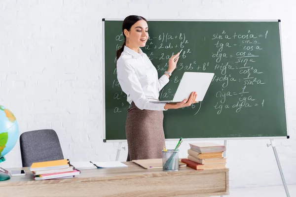 Teacher pointing at chalkboard while holding laptop during online lesson — Stock Photo