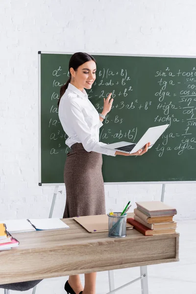 Positiver Lehrer hält Laptop während Online-Unterricht im Klassenzimmer in der Nähe der Tafel — Stockfoto