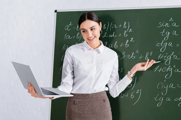 Positive teacher pointing at equation on chalkboard during video chat on laptop — Stock Photo