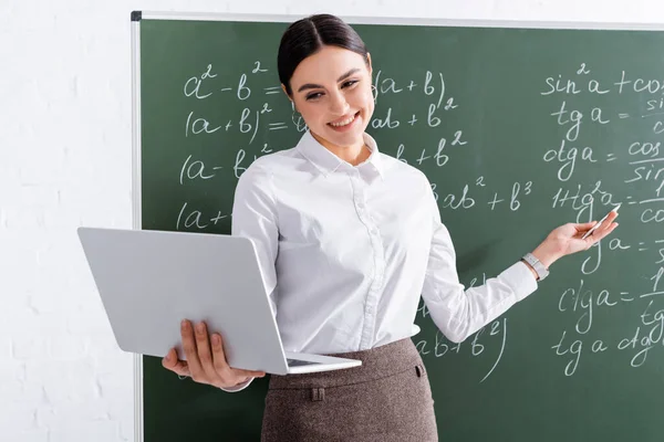 Smiling teacher having video call on laptop near chalkboard in classroom — Stock Photo