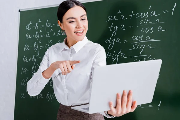 Smiling teacher pointing at laptop during video call near chalkboard in classroom — Stock Photo