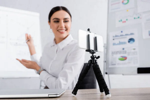 Smartphone on selfie stick near blurred businesswoman having video call near laptop in office — Stock Photo