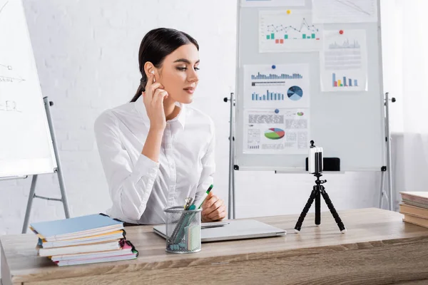 Geschäftsfrau mit Kopfhörer blickt bei Videoanruf im Büro aufs Smartphone — Stockfoto