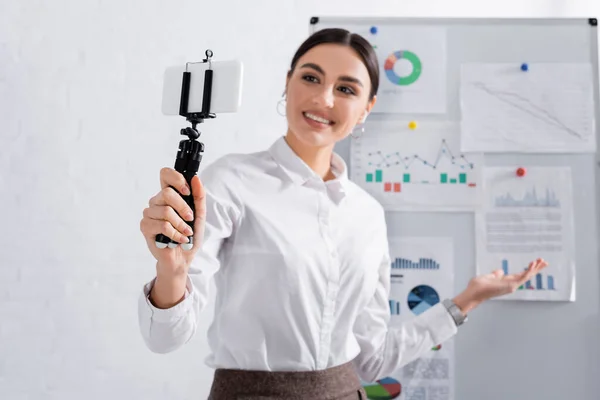 Smartphone en selfie stick en la mano de una mujer de negocios sonriente cerca del rotafolio durante la reunión en línea - foto de stock