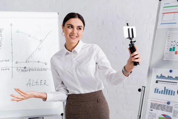 Manager mit Kopfhörer hält Smartphone auf Selfie-Stick bei Online-Meeting in der Nähe von Flipcharts — Stockfoto