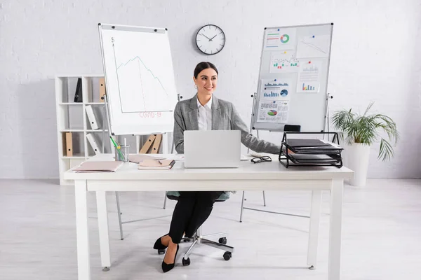 Empresária sorridente sentada perto de laptop e papéis no escritório — Fotografia de Stock