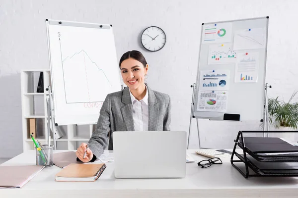 Femme d'affaires souriant à la caméra près de l'ordinateur portable et des papiers dans le bureau — Photo de stock