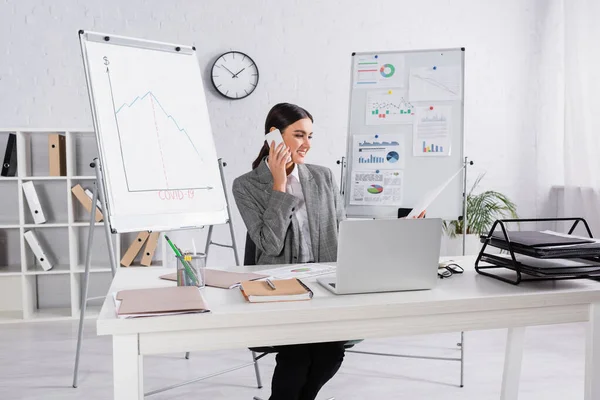 Businesswoman talking on smartphone and holding document near working table in office — Stock Photo