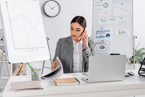 Empresária olhando para papelada e falando em smartphone perto de laptop no escritório — Fotografia de Stock