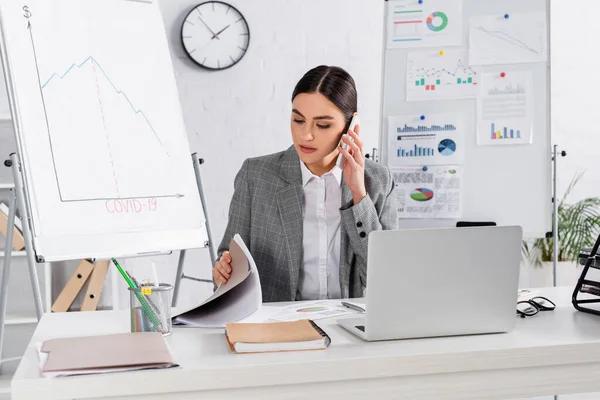 Donna d'affari che guarda i documenti e parla sullo smartphone vicino al laptop in ufficio — Foto stock