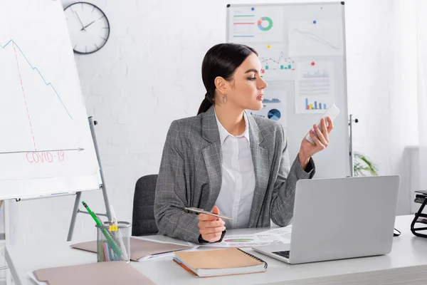 Businesswoman using smartphone and holding pen near laptop and documents — Stock Photo