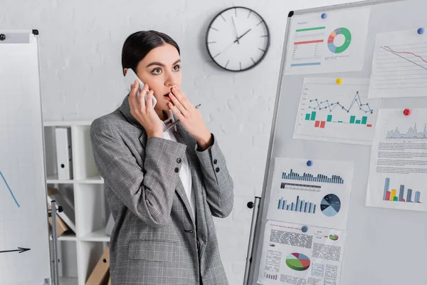 Shocked businesswoman with pen talking on mobile phone near flipchart with graphs — Stock Photo