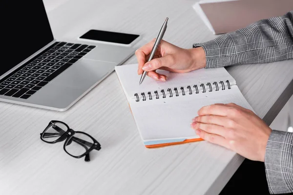 Vista ritagliata della donna d'affari che scrive sul taccuino vicino agli occhiali e ai gadget sfocati — Foto stock