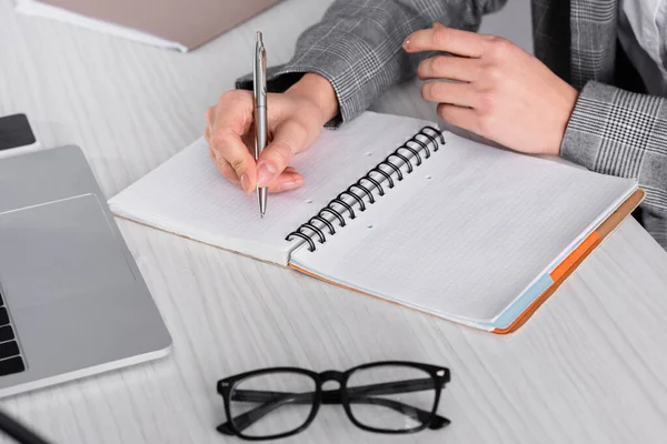 Vista recortada de la mujer de negocios escribiendo en el cuaderno cerca de las gafas y el ordenador portátil - foto de stock