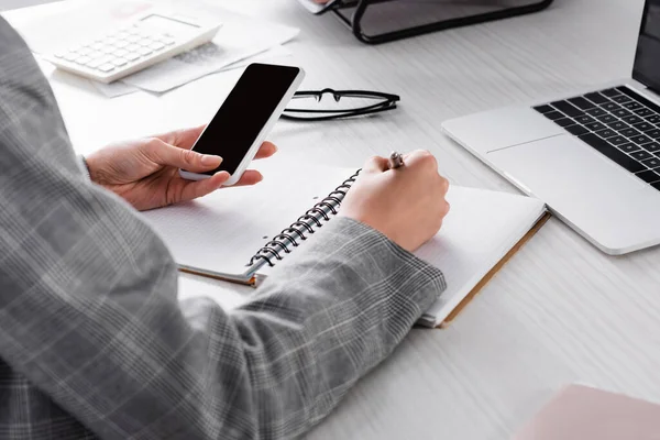 Cropped view of businesswoman with smartphone writing on notebook near laptop and blurred calculator — Stock Photo