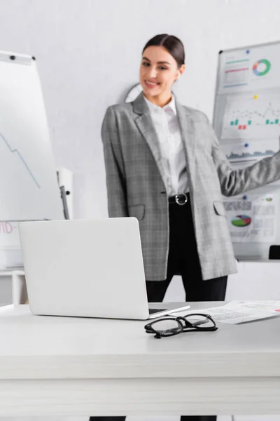 Laptop e óculos perto de mulher de negócios sorridente e flipchart no fundo borrado — Fotografia de Stock