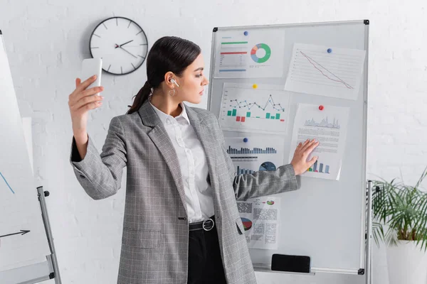 Femme d'affaires dans l'écouteur regardant les documents sur le tableau à feuilles mobiles pendant l'appel vidéo sur smartphone — Stock Photo