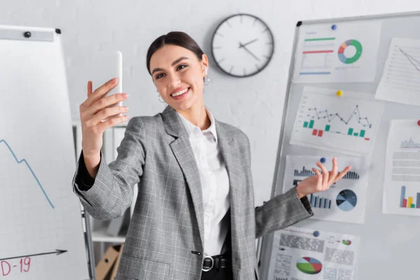 Smartphone en main de femme d'affaires ayant un appel vidéo près de tableaux à feuilles mobiles dans le bureau — Photo de stock