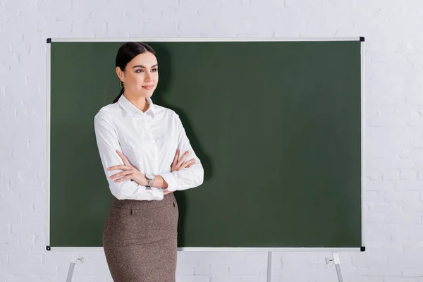 Jeune professeur avec les bras croisés regardant loin près du tableau — Photo de stock