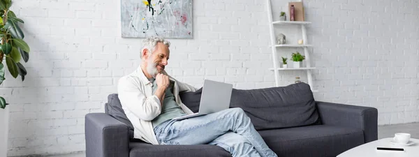 Бородатий чоловік дивиться на ноутбук, сидячи на дивані, банер — стокове фото