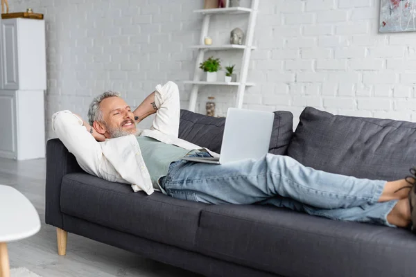 Hombre barbudo feliz mirando el ordenador portátil mientras descansa en el sofá - foto de stock