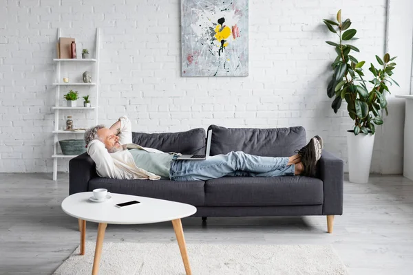 Cheerful bearded man looking at laptop while resting on couch — Stock Photo