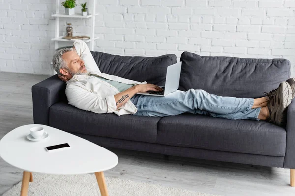 Hombre tatuado usando el ordenador portátil mientras está acostado en el sofá en casa - foto de stock