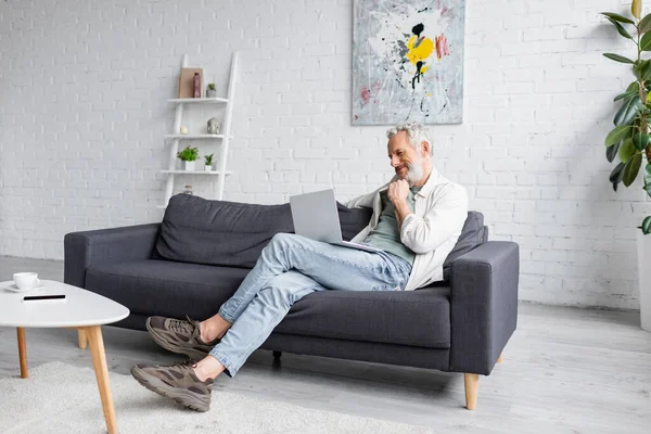 Веселий чоловік використовує ноутбук, сидячи на дивані вдома — стокове фото