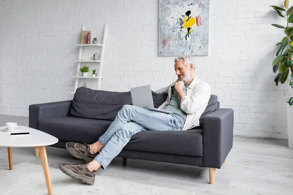 Hombre alegre usando el ordenador portátil mientras está sentado en el sofá en casa - foto de stock