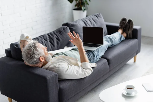 Tattooed man having video call on laptop while lying on couch at home — Stock Photo