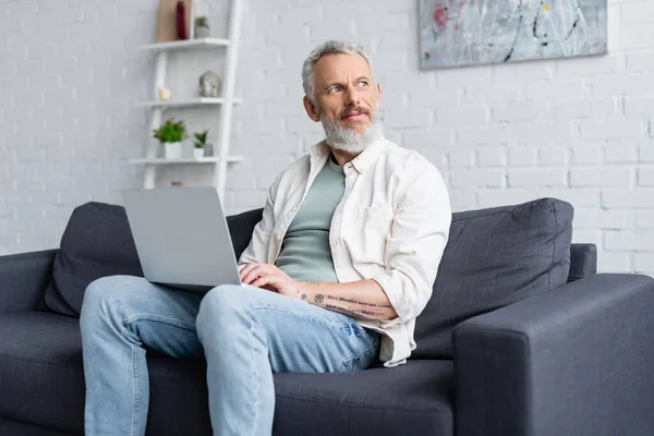 Бородатий чоловік друкує на ноутбуці, сидячи на дивані вдома — стокове фото
