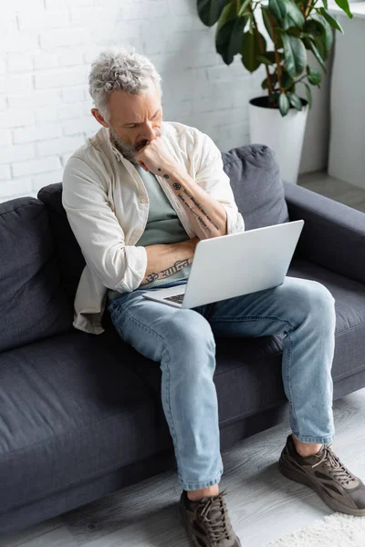 Бородатий чоловік дивиться на ноутбук, сидячи на дивані вдома — стокове фото