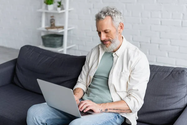 Tätowierter Mann benutzt Laptop, während er zu Hause auf Couch sitzt — Stockfoto