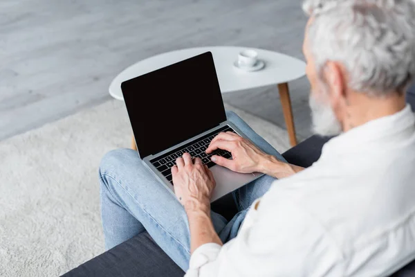 Blurred freelancer with grey hair using laptop while sitting on couch at home — Stock Photo