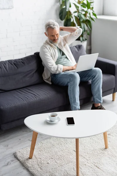 Tazza di caffè e smartphone con schermo bianco sul tavolino vicino all'uomo barbuto utilizzando il computer portatile su sfondo sfocato — Foto stock