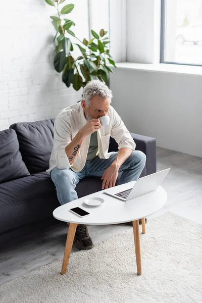 Bearded man drinking coffee and using laptop while sitting on couch near smartphone with blank screen on coffee table — Stock Photo