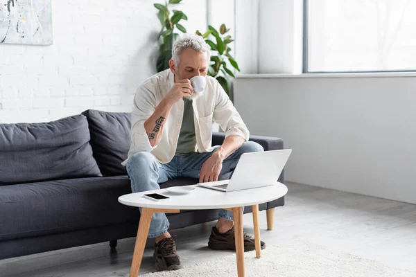 Uomo barbuto bere caffè e utilizzando il computer portatile vicino smartphone con schermo bianco sul tavolino — Foto stock