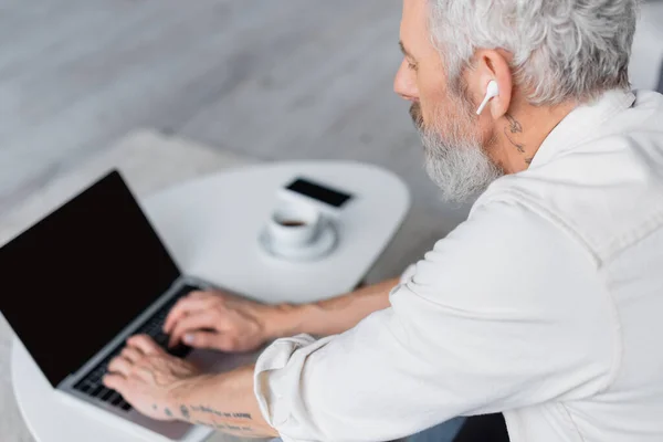 Hombre barbudo en auriculares inalámbricos con ordenador portátil con pantalla en blanco - foto de stock