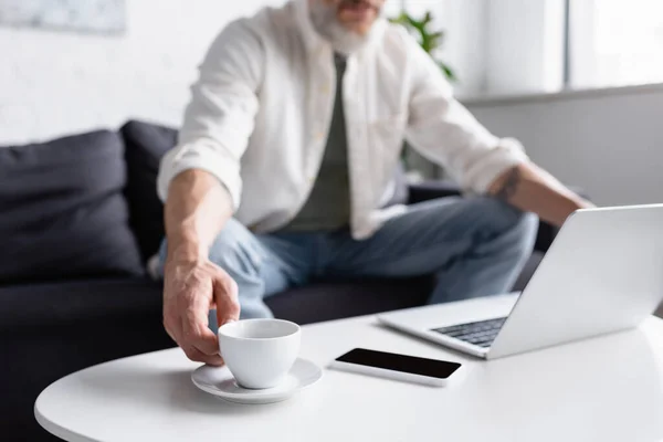 Vista cortada do homem barbudo atingindo xícara de mesa de café perto de gadgets — Stock Photo