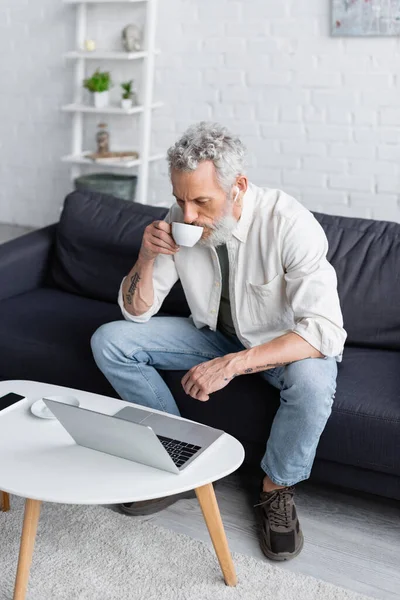 Hombre barbudo en auriculares inalámbricos bebiendo café y mirando el ordenador portátil - foto de stock